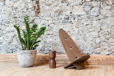 Ein Balanceboard von rollholz in Walnuss zu einem Stuhl zusammengebaut neben einer Faszienrolle aus Holz und einer Pflanze vor einer Steiwand,