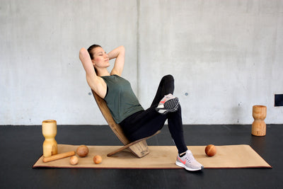 Eine Frau sitzt entspannt auf einem als Stuhl zusammen gebauten Balance Board von rollholz. Neben ihr befinden sich eine Faszienrolle, ein Massagestab und Massagekugeln von Rollholz. Die Unterlage bietet eine Korkmatte