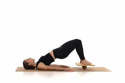 Eine Frau liegt mit dem Rücken auf einer Korkmatte. Ihre Füße stehen auf einem Brett, das auf einer Massagekugel von rollholz balanciert wird.