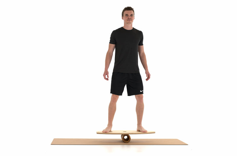 Ein Mann steht auf einem Balance Board, das auf einer Faszienrolle aus Holz aufliegt. Die Unterlage bietet eine Korkmatte von Rollholz.