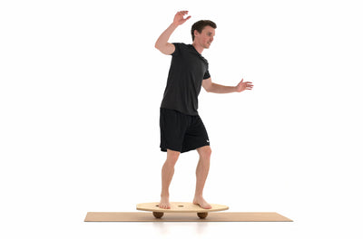 Ein Mann steht auf einem Balance Board von rollholz, das auf 2 Massagekugeln aufliegt und flattert mit den Armen. Die Unterlage bietet eine Korkmatte.