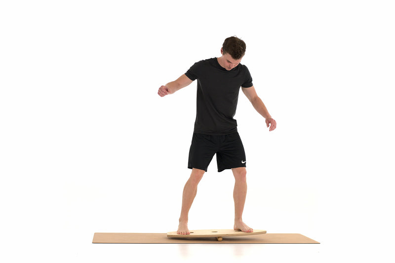 Ein Mann steht auf einem Balance Board, das auf einem Massagestab liegt. Als Unterlage dient eine Korkmatte von rollholz