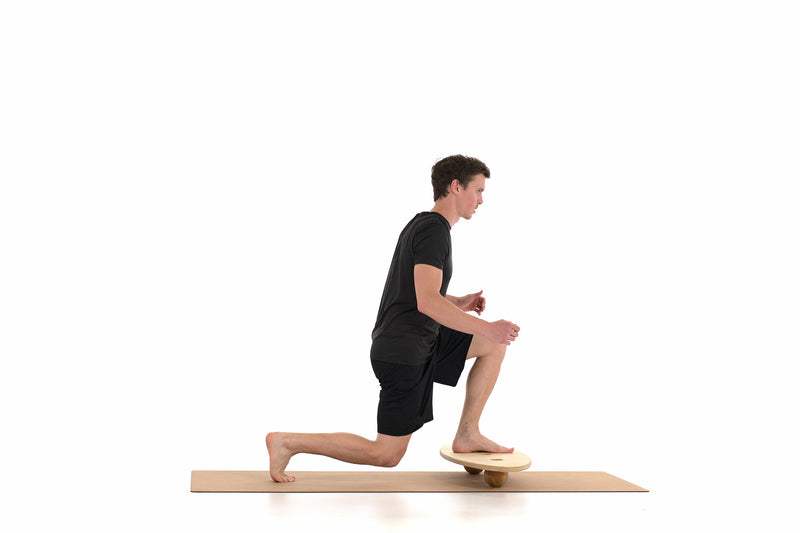 Ein Mann steht mit einem angewinkelten Bein auf dem Boden, der andere Fuß steht auf einem Balance Board von rollholz, das auf 2 Massagekugen aufliegt. Die Unterlage bietet eine Korkmatte